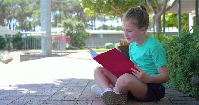 Child girl sitting on brick paving in schoolyard, reading red notebook in teal tee, copy space
