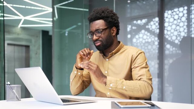Sick African American businessman coughs and holds hand to chest at desk. Unwell worker sitting at corporate office feels pain from bad cold or flu virus symptoms during workday indoors.