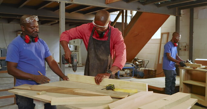Three African American coworkers operating cordless drill and marking wooden planks at workshop