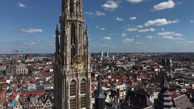 Golden clock faces adorn gothic Antwerp cathedral tower up close, historic rooftops and distant city skyline spreading beneath scattered clouds near ornate church spires. Aerial drone orbit shot