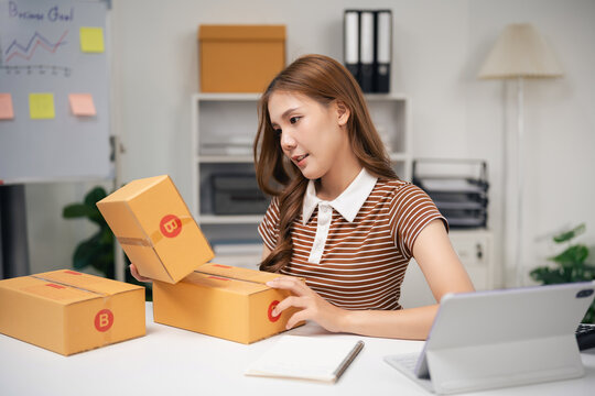 Young woman preparing and organizing cardboard delivery box for online business at home office workspace with tablet and notepad