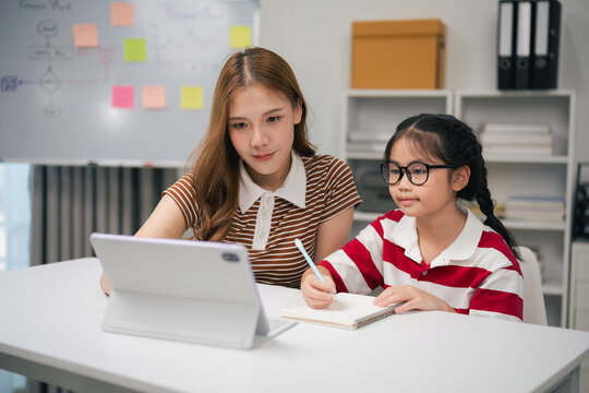 Young woman helping girl with homework using digital tablet at modern study desk in home interior