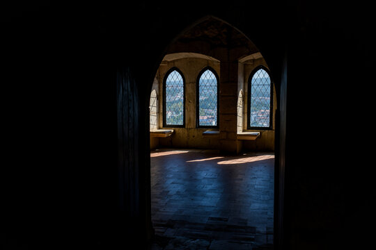 Dark Gothic stone interior seen through a pointed arch with three leaded glass windows illuminated by sunlight