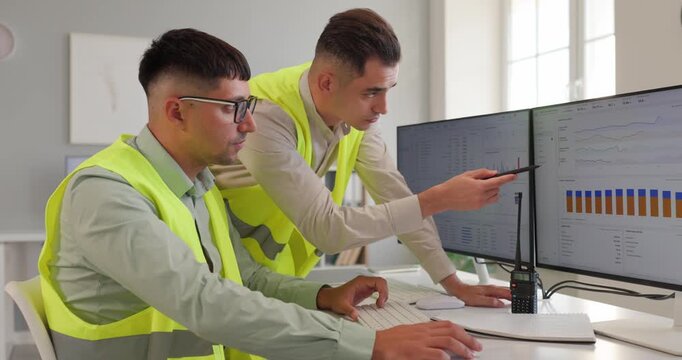Power plant operators monitor energy generation dashboard. Two engineers in safety vests discuss production charts with a walkie talkie on the desk. Teamwork and control in a power plant.