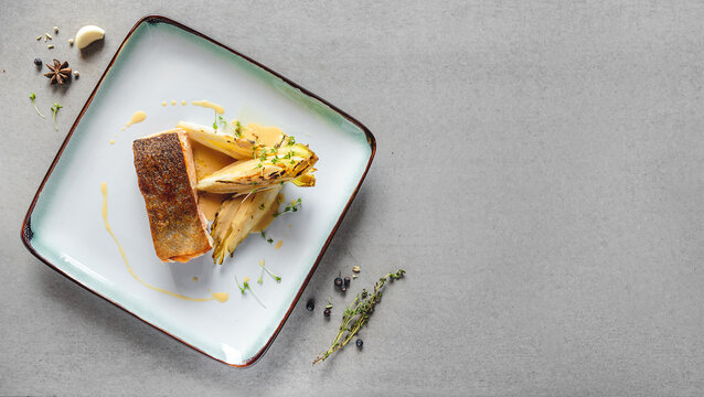 Pan seared salmon fillet with charred endive and creamy sauce on square white ceramic plate positioned on gray concrete tabletop for restaurant menu