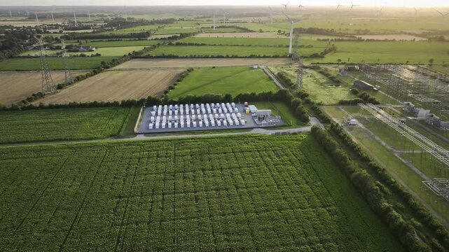 Aerial footage of grid battery energy storage system (BESS) facility located next to an electrical substation and wind turbines in a rural agricultural landscape at sunset.