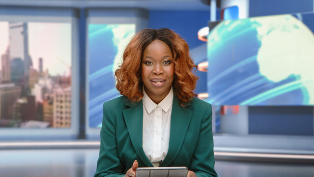 Portrait of a Black Female News Anchor Presenter Holding a Tablet and Reporting Top Headlines to Camera at the Anchor Presenter Desk, Hosting a Live Evening Television News Broadcast From the Studio.
