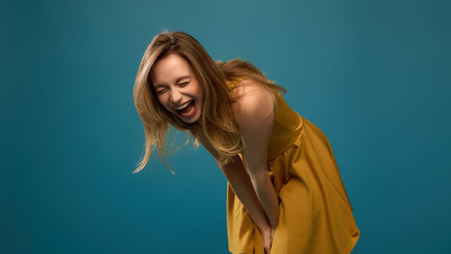 Joyful woman bending forward with laughter against teal studio background in mustard dress. Positive wellbeing, lifestyle editorial, casual fashion concept.