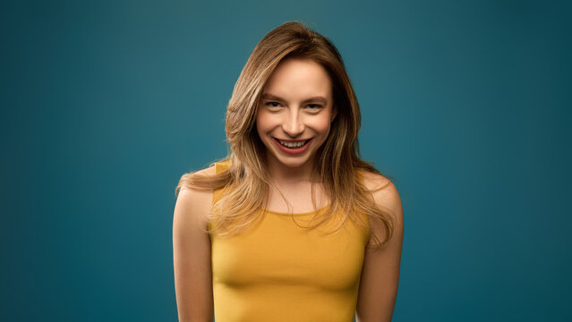 Smiling young woman looking at camera with playful confidence against teal studio background. Personal branding, fashion retail, positive lifestyle concept.