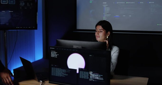 Young Asian woman reviews computer screens as Asian male colleague leans closer and explains software interface, supporting system analysis, technology collaboration, cybersecurity workflow.