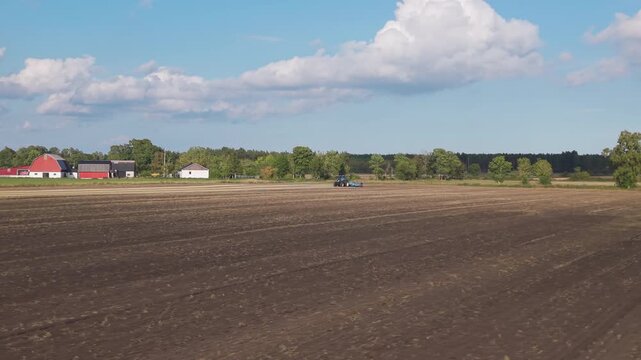 A distant tractor with tillage equipment is visible across a broad stubble field, with red barns, trees and white clouds against a blue sky.