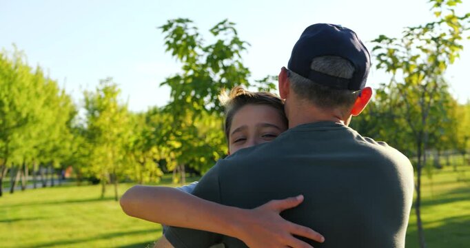 Father embraces son in sunny park. Boy and dad share tender reunion outdoors during summer. Family love, support and connection create warm lifestyle moment for bonding, childhood and parenting.