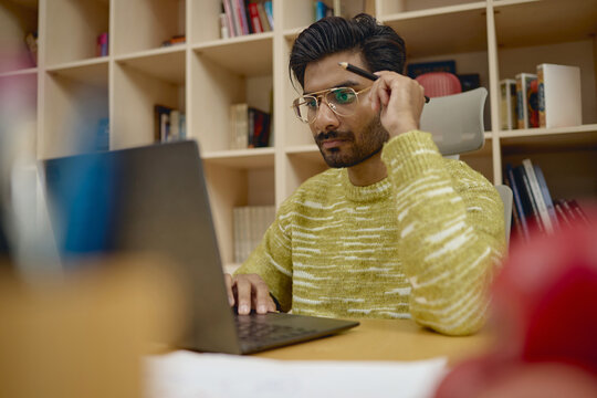 Young adult South Asian man working on laptop in modern office, thinking and holding pencil. Visual concept for remote education, creative business, software development advertising