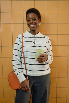 Portrait of smiling young adult African American woman holding reusable coffee cup indoors. Concept of eco friendly lifestyle and modern urban commuting for marketing campaign, vertical