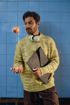 Vertical shot of young adult South Asian man holding laptop case and tossing apple during work break. Image suitable for modern education and career campaigns