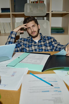 Vertical portrait of young adult Caucasian man sitting at office desk with documents, looking at camera with tired expression. Suitable for corporate burnout, workload stress, deadline concept