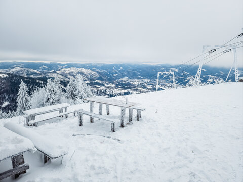 Snow-covered wooden picnic tables and benches on a mountain viewpoint during winter