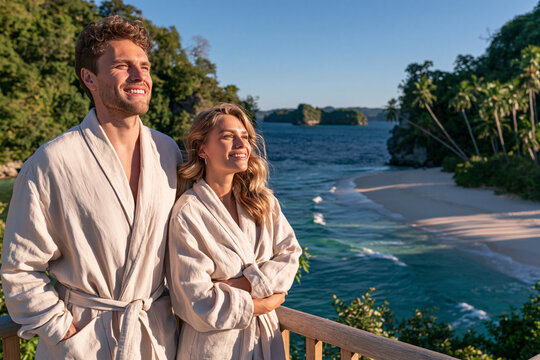 A happy couple in white bathrobes stands on a wooden balcony overlooking a tropical beach and ocean at sunset enjoying the serene coastal view and warm golden light