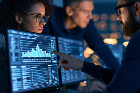 A diverse team of data analysts collaborates late at night examining intricate financial charts and software code on multiple glowing monitors in a modern professional office environment