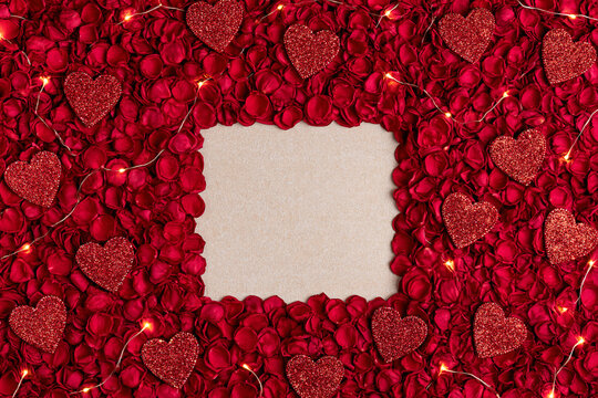 A top down view of a rectangular beige paper blank surrounded by lush red rose petals glittering heart ornaments and twinkling fairy lights creating a romantic and festive atmosphere
