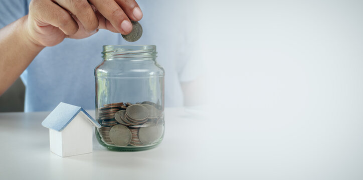 Businessman hand putting coin into glass jar next to wooden house model on white desk, concept of house savings, home loan planning, real estate investment, and future retirement fund.