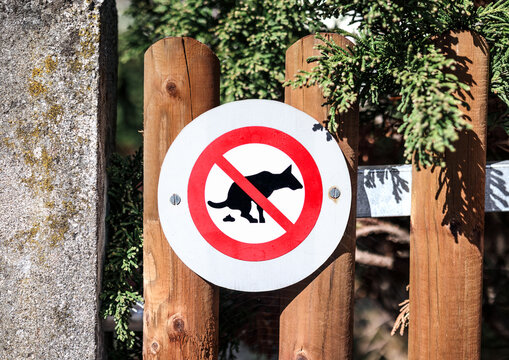 No dog pooping sign on fence. Dog fouling prohibited graphic mounted on fence. Round sign with a dog defecating inside a red circle with a red diagonal prohibition slash. Selective focus.