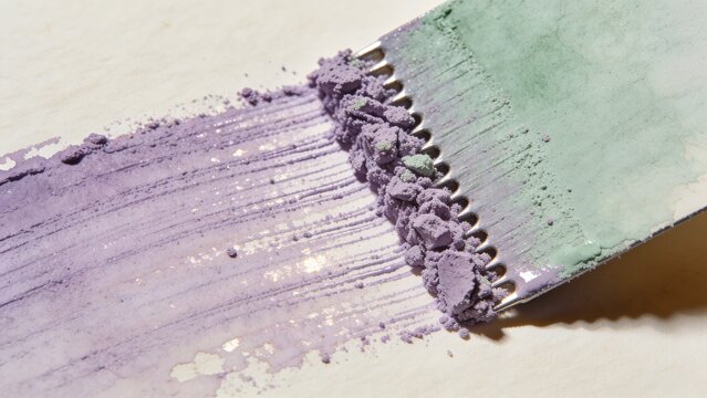 Scraping notched metal tool dragging mint and lavender pigment across white paper leaving mica