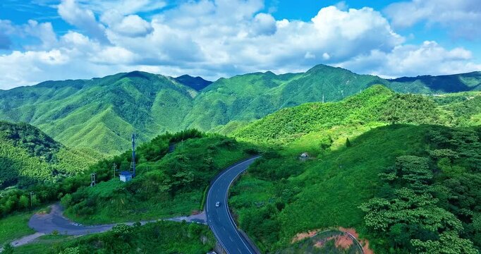 Winding asphalt road snakes through lush green Siming Mountain under a bright blue sky with fluffy white clouds in Ningbo.