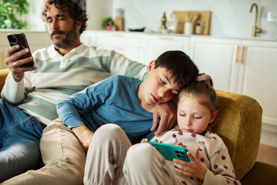 Siblings watch smartphone on couch while dad scrolls