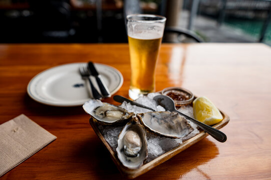Fresh oysters placed on ice with a lemon slice and dipping sauce beside a beer on a wooden table