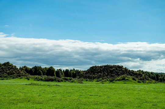 Green field stretches under a blue sky filled with clouds, with rolling hills and trees