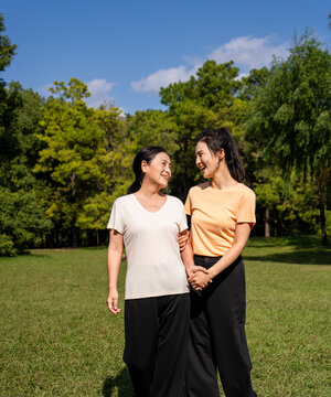  Happy Asian senior mother and adult daughter holding hands, smiling together in green park on sunny day, family bonding outdoor portrait