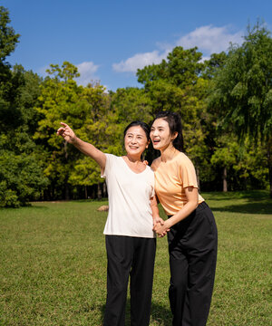  Happy Asian senior mother and adult daughter holding hands, smiling together in green park on sunny day, family bonding outdoor portrait