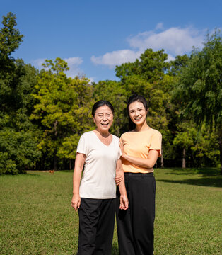  Happy Asian senior mother and adult daughter holding hands, smiling together in green park on sunny day, family bonding outdoor portrait