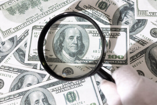 Gloved Hand holding a magnifying glass inspecting a bunch of US dollar bills. Financial investigation tracing auditing concept