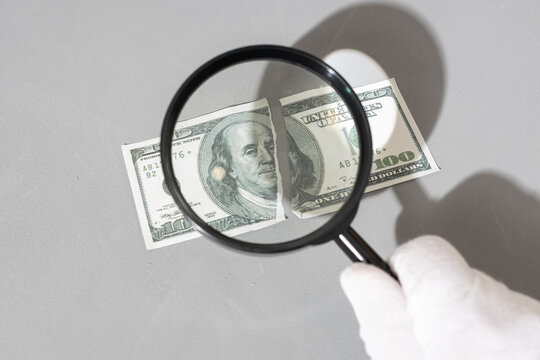 hand holding magnifying glass inspecting torn one hundred dollar banknote