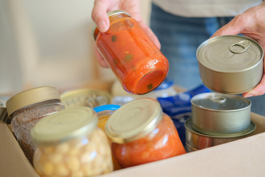 Woman hands placing canned and jarred groceries into cardboard donation box. Food bank collection for families in need. Community charity and food aid. Volunteering and giving concept.