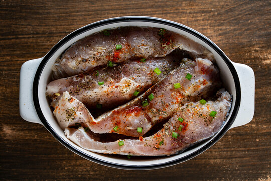 Raw fish steaks marinated with spices in baking dish top view, closeup. Fresh raw fish pieces seasoned with herbs, paprika, and green onions in ceramic baking bowl on dark wooden table