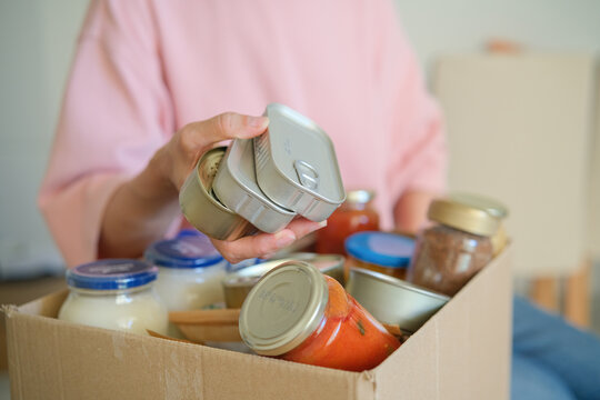 Person placing canned food into cardboard donation box filled with groceries. Food bank collection and community support. Food donation, hunger relief and charitable giving.
