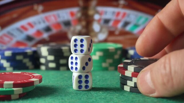 Hand stacking poker chips beside tower of dice on green casino table with roulette wheel in background, close up gambling concept and tabletop game scene
