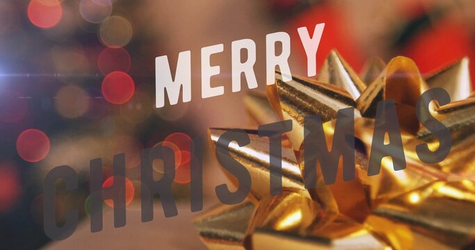 Large gold gift bow resting near MERRY CHRISTMAS text against blurred red lights, copy space