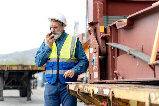 Experienced logistics inspector coordinating transport operations using a walkie talkie near a container chassis structure.
