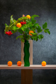 Sprig of red guelder-rose (viburnum) in a green vase on a wooden shelf, accompanied by orange spheres.