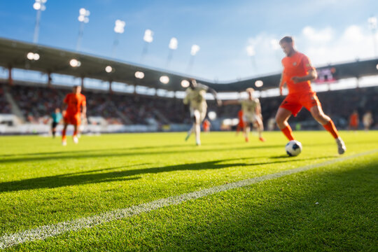 Football player in orange kit dribbling the ball on a sunlit green pitch during a match in a stadium