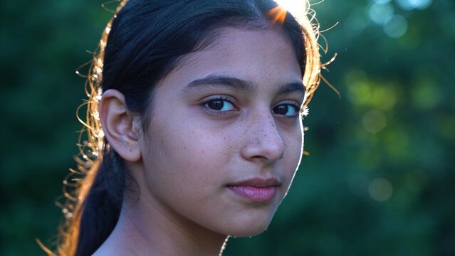 Teenage girl with dark hair and freckles facing viewer near blurred green foliage, AI-generated
