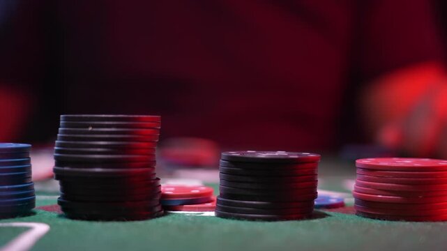 Close-up of poker chips stacked on green felt table. Black red and blue chips form neat rows. Red lighting highlights glossy surfaces. Background blur creates casino mood