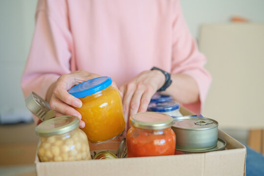 Woman volunteer placing canned and jarred groceries into donation box. Food bank collection for families in need. Community charity and food aid