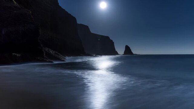 Moonlit ocean waves wash ashore beside rocky cliffs, a sandy beach, and an offshore sea stack. Nature documentaries, travel promotion, and coastal tourism campaigns.