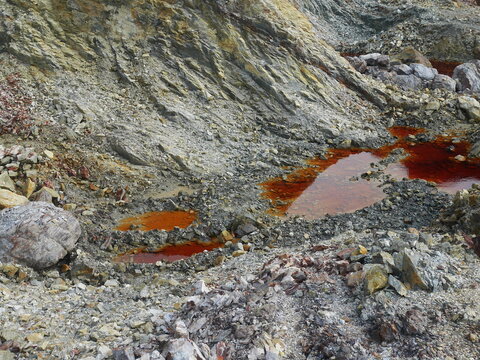 An expansive rocky slope covered with fractured gray stones and scattered mineral-stained pools. Orange and reddish water traces wind through the rugged terrain, contrasting with the layered rock.