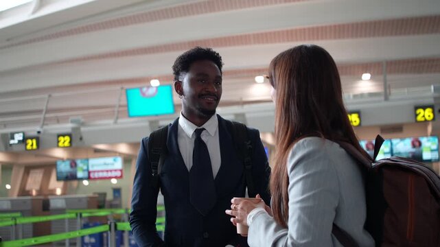Multiethnic business couple with backpacks talking and smiling while waiting for their flight at the check in counters in the airport terminal, discussing their upcoming business trip